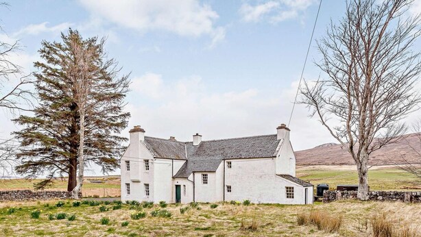 Un bâtiment blanc avec un mur de pierre, une cheminée en pierre et un champ verdoyant avec un arbre.