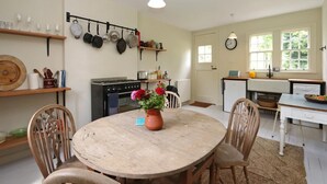 Une cuisine équipée d’une cuisinière, d’un four et de casseroles suspendues. On y trouve une table ronde en bois, entourée de chaises, et un vase rempli de fleurs.