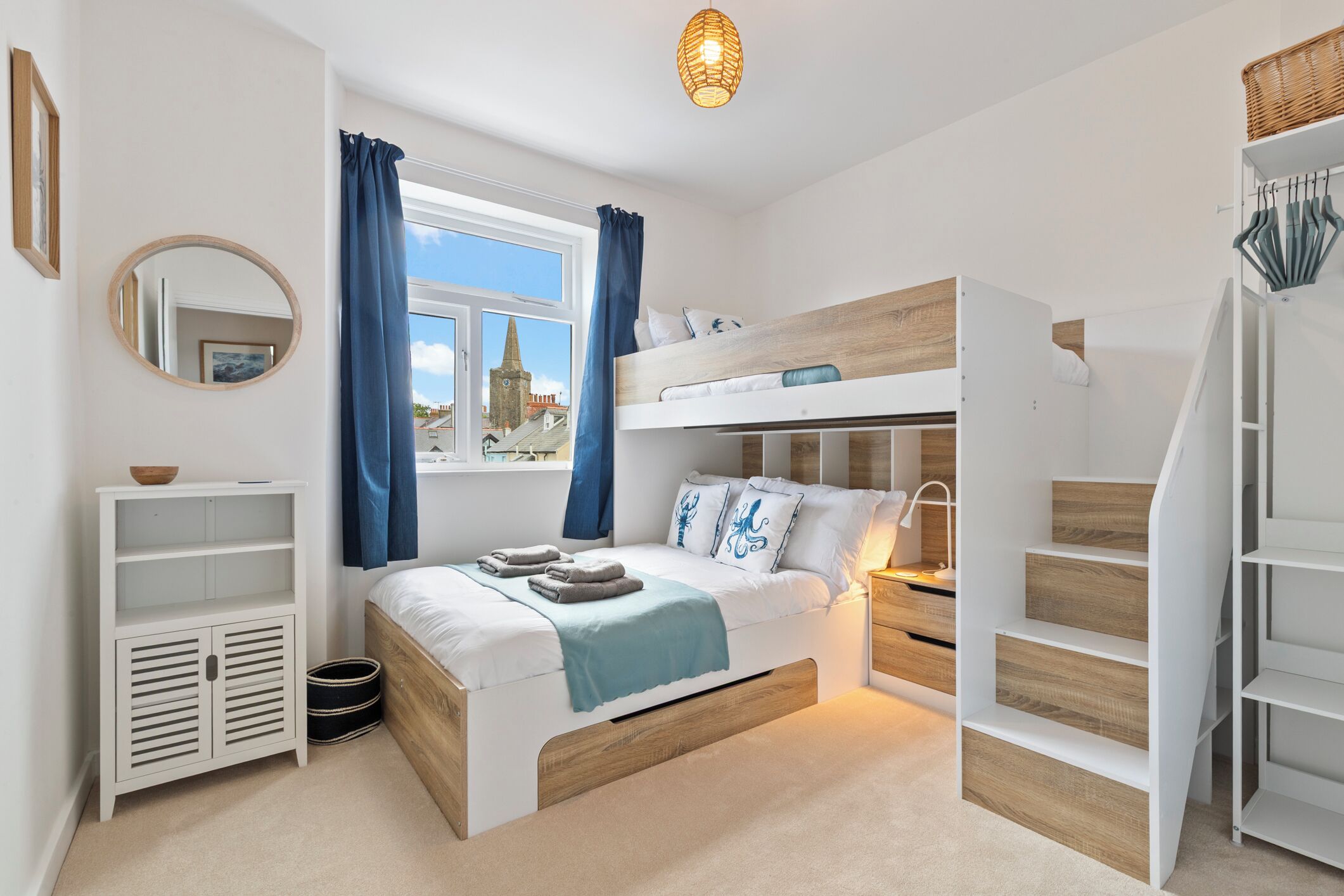 Une chambre avec un lit superposé, une table de chevet, une lampe, une fenêtre avec des rideaux, un miroir et un petit meuble.