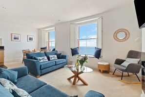 Ein geräumiges Wohnzimmer mit einem blauen Sofa, einem Glastisch und Meerblick durch große Fenster.