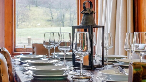 Una mesa de comedor servida con platos, vasos y cubiertos, una vela en un farol, y una vista de árboles a través de una ventana.