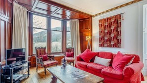 Un salón acogedor con un sofá rojo, una mesa de centro de madera y un televisor sobre un mueble bajo. Amplias ventanas ofrecen vistas al exterior.