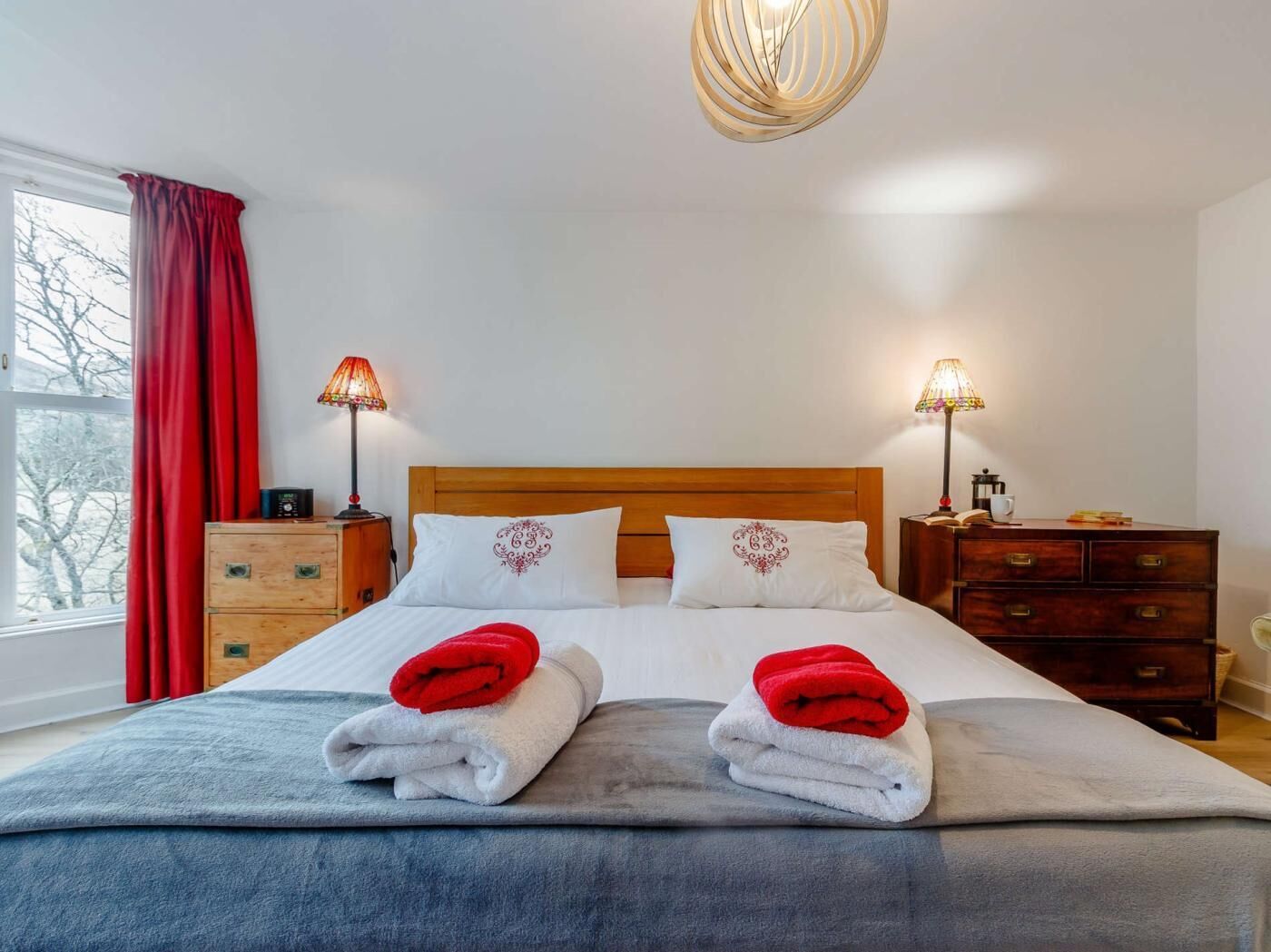 A neatly made bed with two pillows, a wooden headboard, two bedside tables with lamps, a dresser, and a window with red curtains.