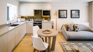A modern kitchen with white cabinets, a dining area with a round table and chairs, a grey sofa with patterned cushions, and two framed pictures on the wall. - 1 Bed in Windermere (oc-lr229) (Windermere)