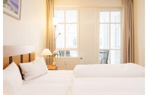 A hotel room with two beds, a nightstand with a lamp, a window with curtains, and a radiator. - Ferienwohnung mit Balkon und Freizeitangeboten in Sellin (Sellin)
