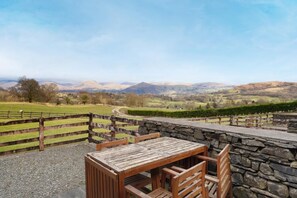 A rustic outdoor seating area with a stone wall and wooden table and chairs, overlooking a scenic valley with hills and a clear sky. - 3 Bed in Hawkshead (oc-lr238) (Hawkshead Hill)