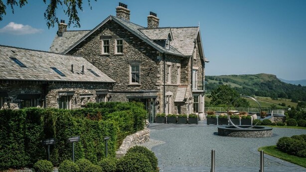 A stone building with a gravel driveway, a circular water feature, and a view of green hills in the background. - 1 Bed in Windermere (oc-lr232) (Windermere)