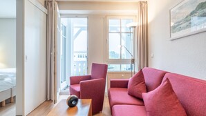 A modern hotel room with a red sofa, a wooden side table, a lamp, and a large window with a view. - Ferienwohnung Seepark Sellin mit Ahoi Badewelt & Balkon (Sellin)