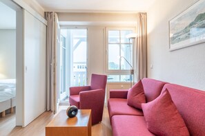 A modern hotel room with a red sofa, a wooden side table, a lamp, and a large window with a view. - Ferienwohnung Seepark Sellin mit Ahoi Badewelt & Balkon (Sellin)