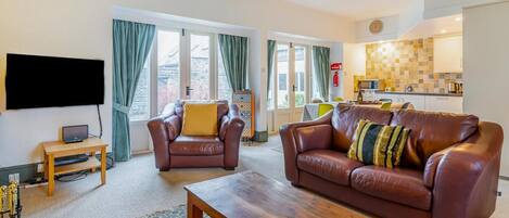 A living room with a sofa, armchair, coffee table, TV, and a kitchen area with a tiled backsplash.