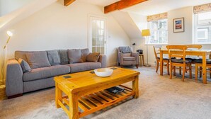 A cozy living room with a sofa, a wooden coffee table, a dining table with chairs, a lamp, and a window with curtains. - 2 Bed in Ambleside (oc-lr041) (Ambleside)