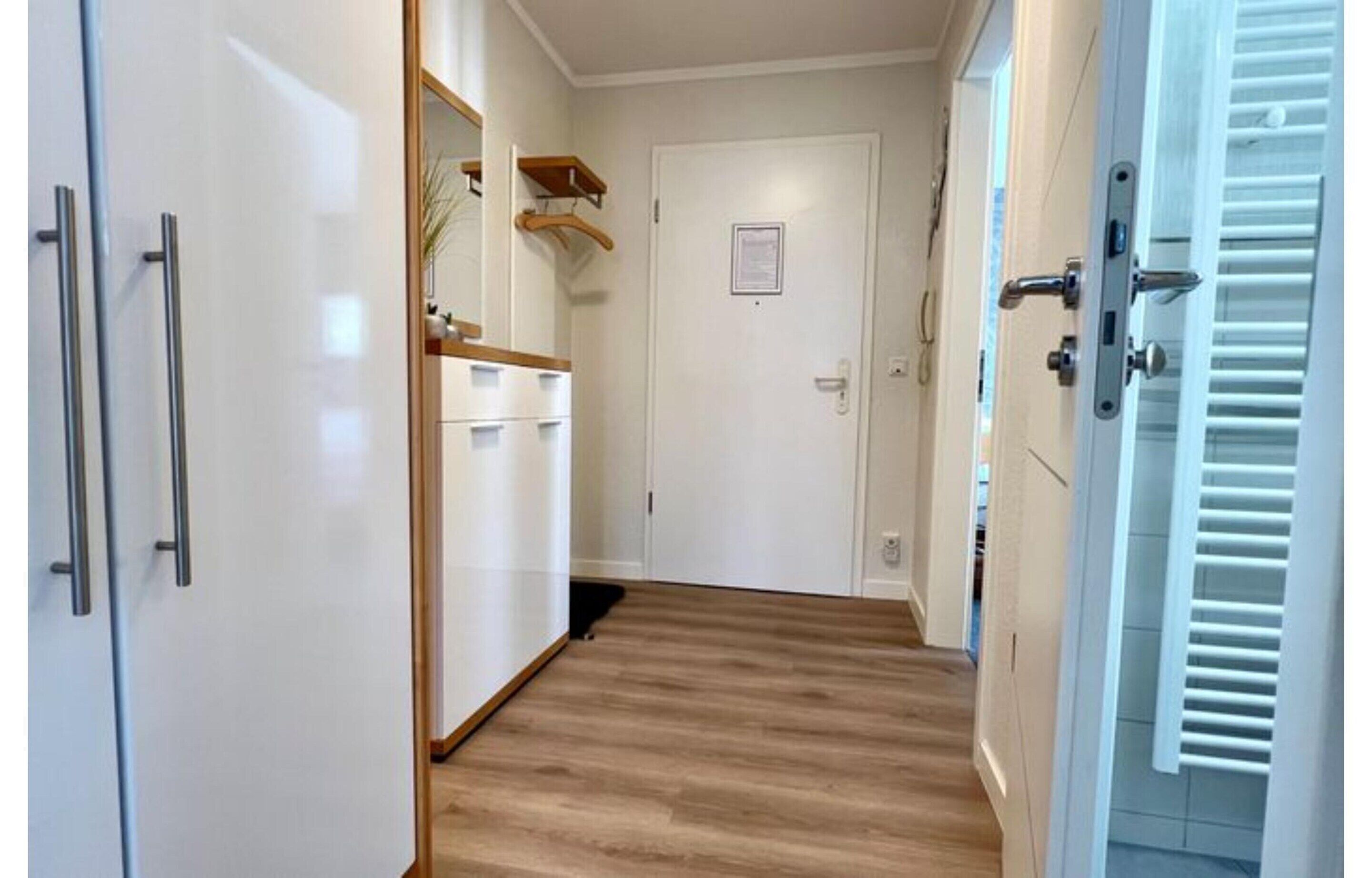 A hallway with a wooden floor, white walls, a door with a small sign, and a white door with a handle.