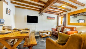 Een rustieke kamer met zichtbare houten balken, een houten eettafel met kommen, een bank en een aan de muur bevestigde televisie.