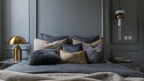 Un lit avec une couverture grise, plusieurs oreillers, une lampe dorée et un luminaire fixé au mur.