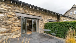 A stone building with a glass door entrance, a patio with a table and chairs, and a hedge in the background. - 1 Bed in Windermere (oc-lr225) (Windermere)