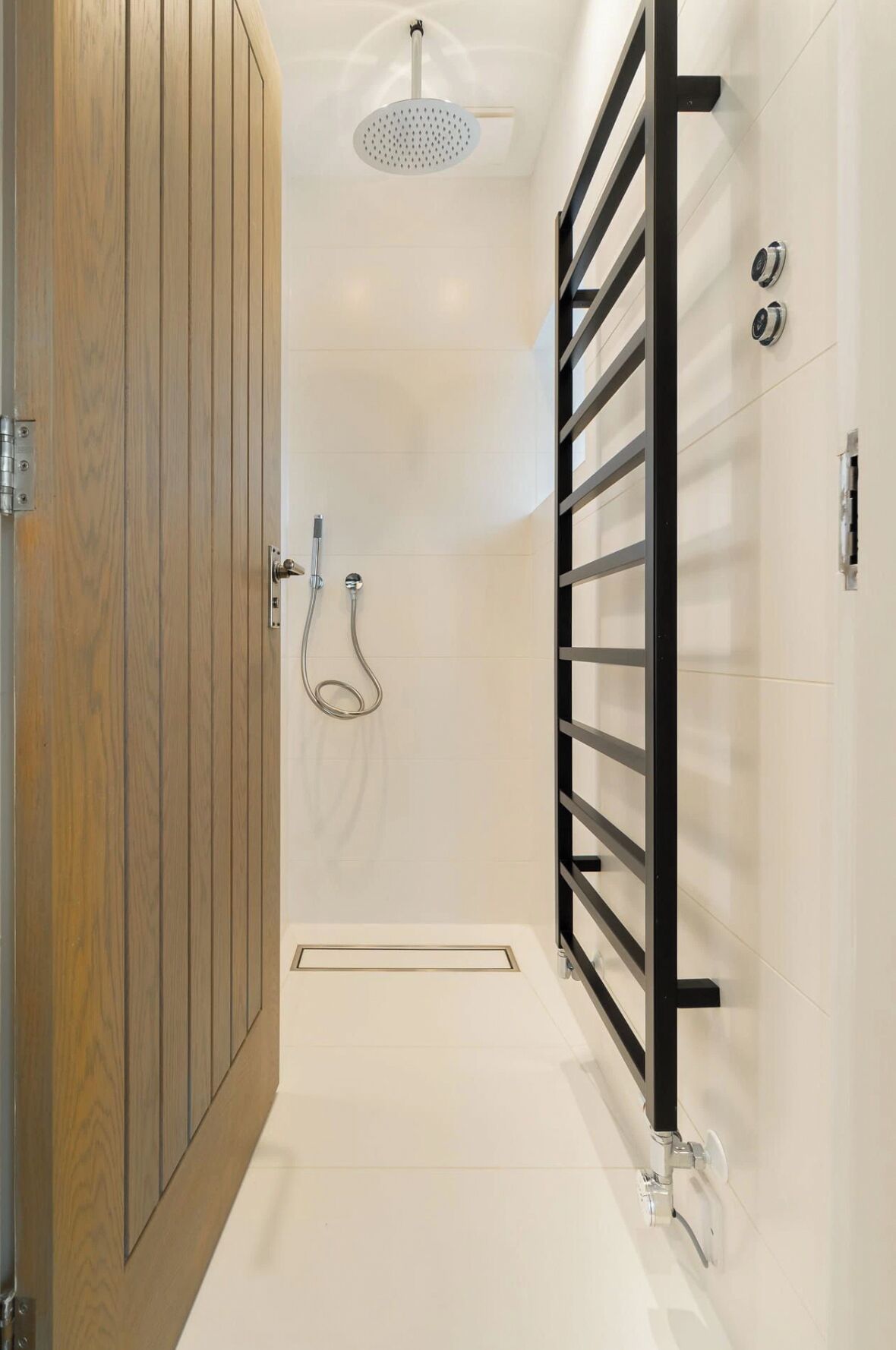 A modern bathroom with a wooden door, a shower with a rain showerhead, a black towel rack, and a white tiled floor.