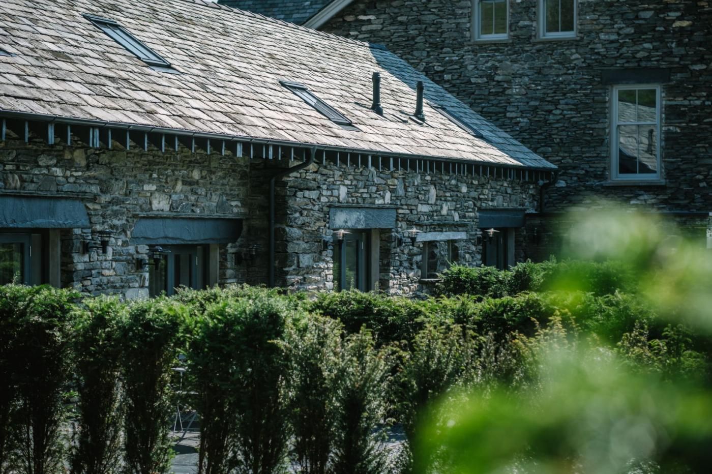 A stone building with a sloped roof, multiple windows, and a well-maintained hedge in the foreground.