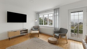 Een woonkamer met een flatscreen televisie, twee fauteuils, een houten vloer, een groot raam en uitzicht op de omgeving.