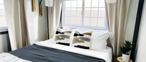 A neatly made bed with a dark-colored bedspread and multiple pillows. There is a small bedside table with a plant on it. The room has a window with curtains and a view of the outside.