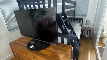 A bedroom with a bunk bed, a wooden dresser with a flat-screen TV, a bedside table with a lamp, and a window with blinds.