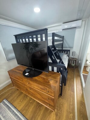 Une chambre avec un lit superposé, une commode en bois sur laquelle est posée une télévision à écran plat, une table de chevet avec une lampe et une fenêtre avec des stores.