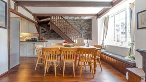 Ein Essbereich mit Holzbänken und einem Tisch, im Hintergrund eine Küche, eine Steinwand und eine Treppe.