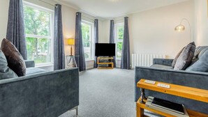 Un salon moderne avec des canapés gris, un téléviseur à écran plat, une lampe et une petite table d’appoint en bois.