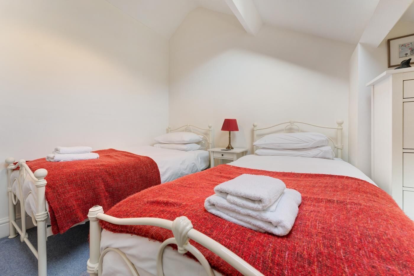 Deux lits simples avec des couvertures rouges et des oreillers blancs, une table de chevet avec une lampe rouge et une armoire blanche.