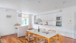 A modern kitchen with white cabinets, a dining table with chairs, and a large window. - 3 Bed in Ambleside (oc-lr223) (Claife)