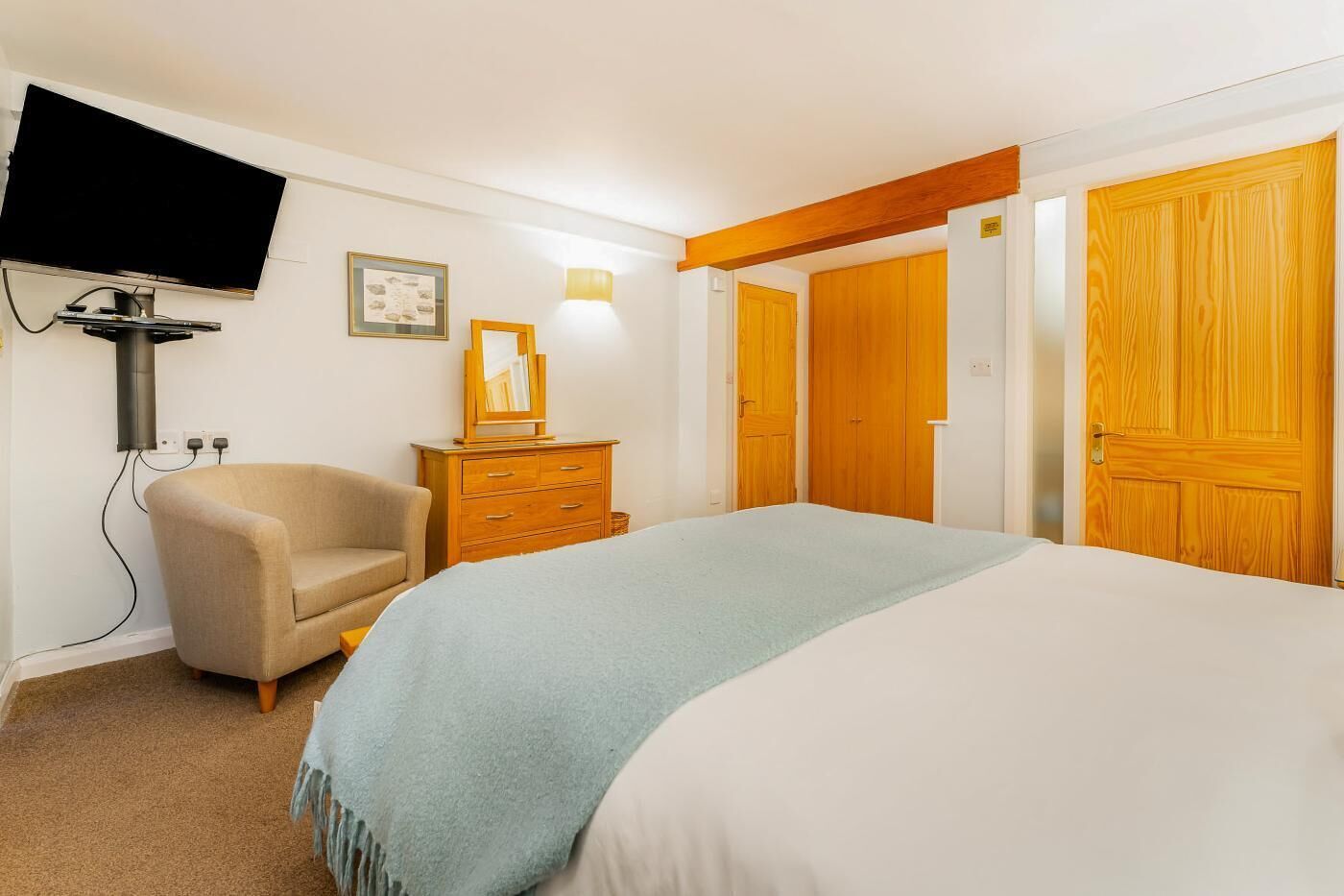 Un dormitorio con cama, una silla, cómoda y televisor montado en la pared, además de puertas de madera.