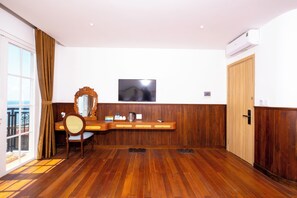Quarto de hotel com piso de madeira, televisão presa na parede, armário de madeira, cadeira com assento trançado e uma janela grande com cortinas.