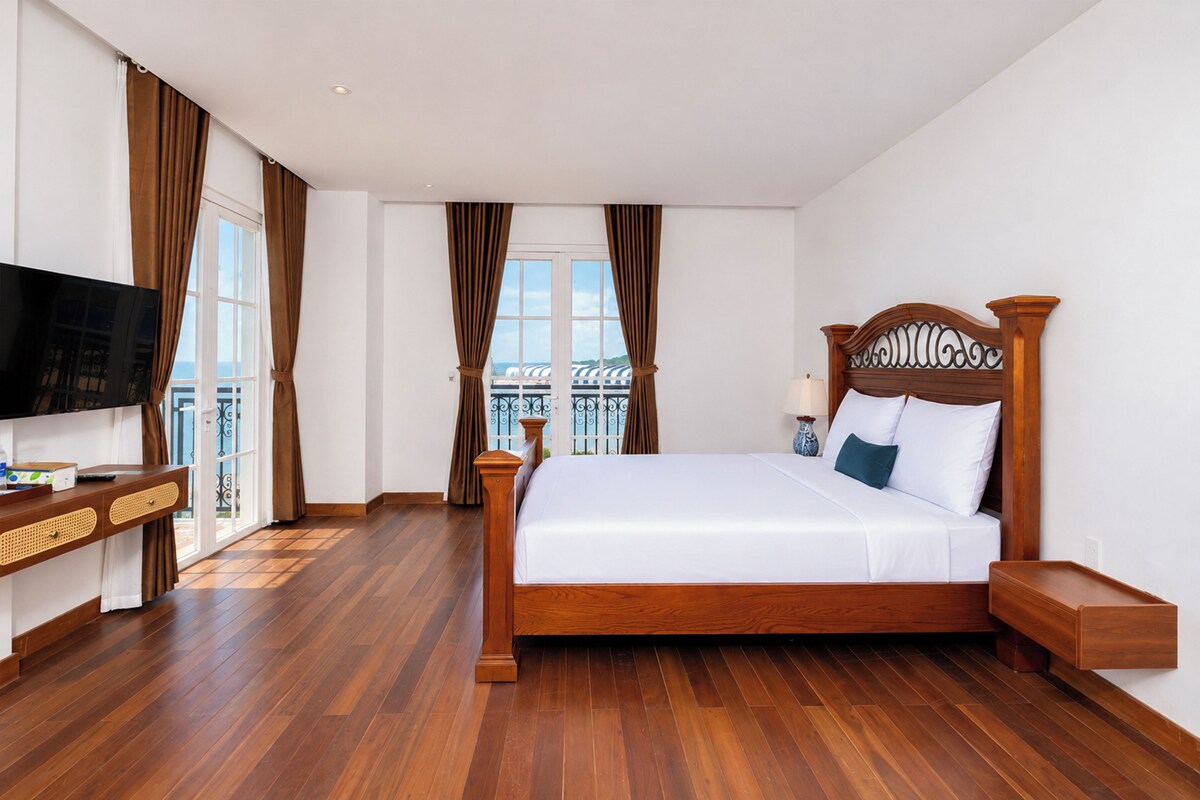 Habitación de hotel con una cama grande, cabecera de madera, un televisor en un soporte, una mesita de noche y vistas al balcón exterior.