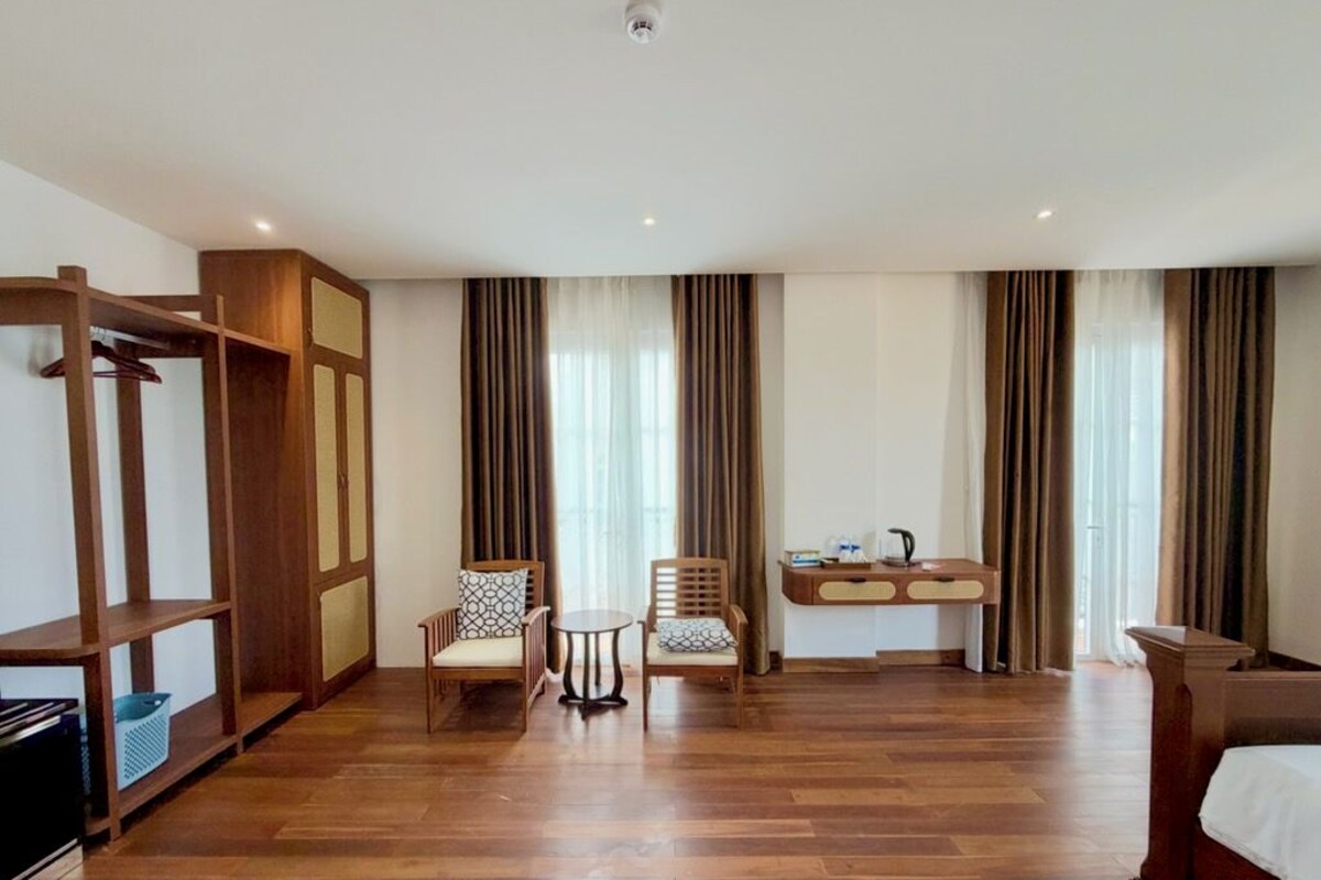 Habitación de hotel con piso de madera, dos sillas, una mesita, un escritorio con tetera y una cama.