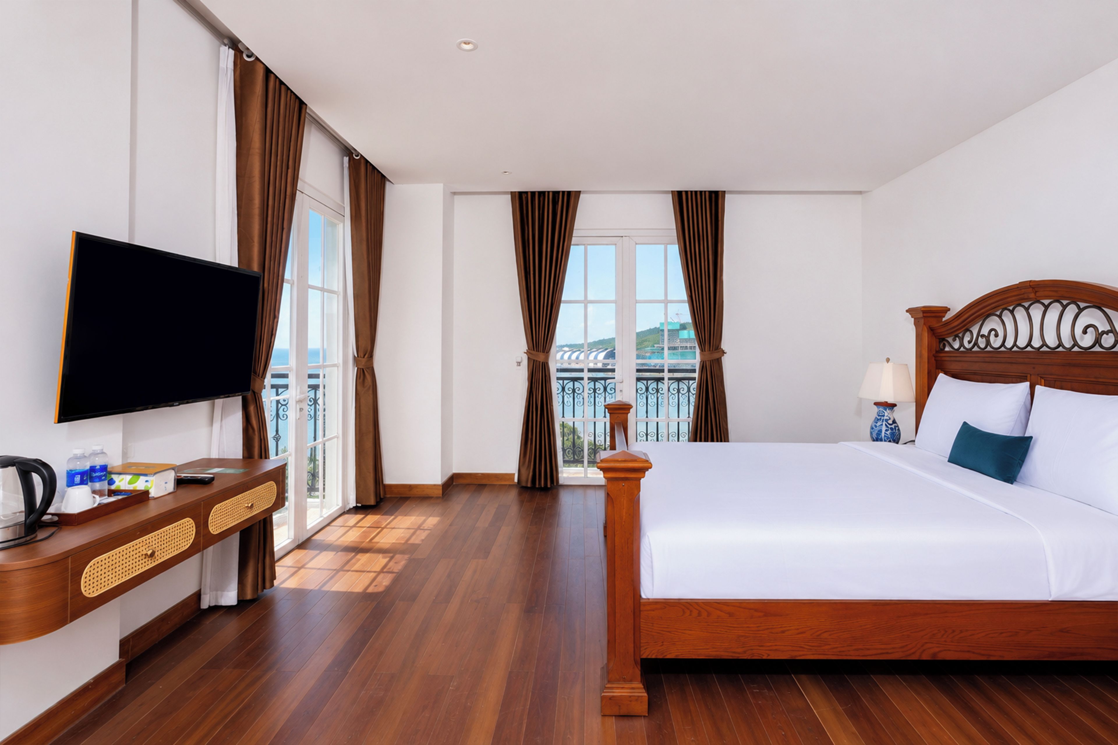 A hotel room with a large bed, a flat-screen TV, a wooden dresser, and a view of the cityscape through the window.