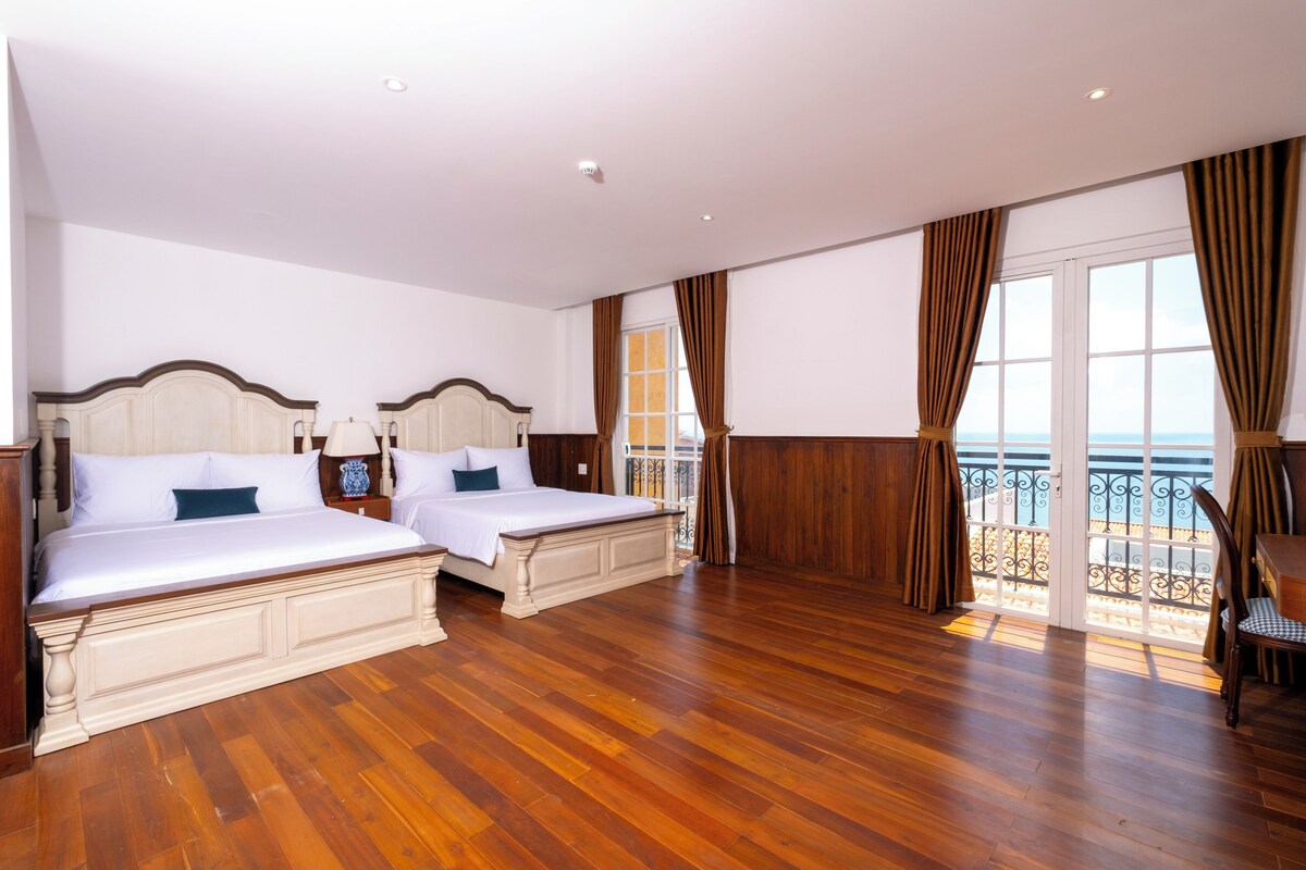 Habitación de hotel con dos camas, piso de madera, un balcón con vistas y un ventanal grande con cortinas.