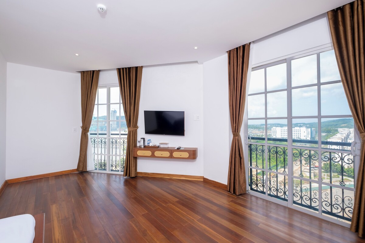 Ein geräumiges Zimmer mit großem Fenster, einem wandmontierten Flachbildfernseher und einem Holztischchen mit verschiedenen Gegenständen.