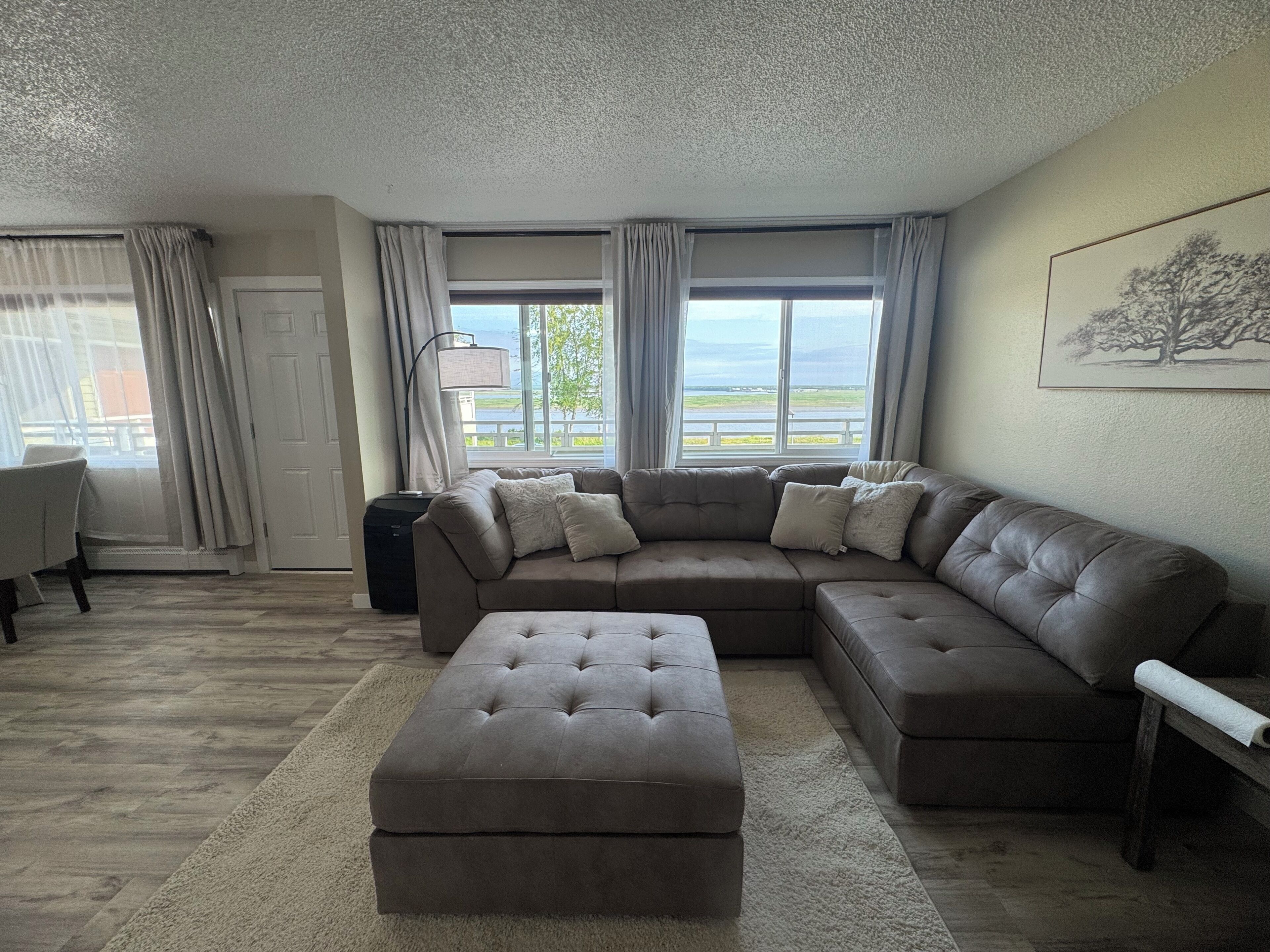 Una sala de estar con un sofá seccional, un reposapiés, una lámpara y vistas al exterior.