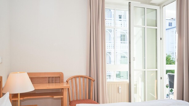 A hotel room with a bed, a desk with a lamp, a chair, and a large window with curtains. - Charmante Ferienwohnung fur 3 mit Balkon im Seepark Sellin (Sellin)
