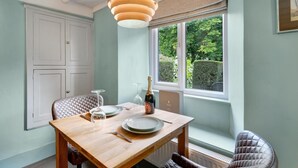 A dining area with a wooden table, chairs, a bottle of champagne, and a window with a view of greenery. - 1 Bed in Bowness On Windermere (oc-lr171) (Bowness on Windermere)