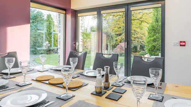 Ein Essbereich mit Tisch für vier Personen, großen Fenstern und Blick auf einen Garten.