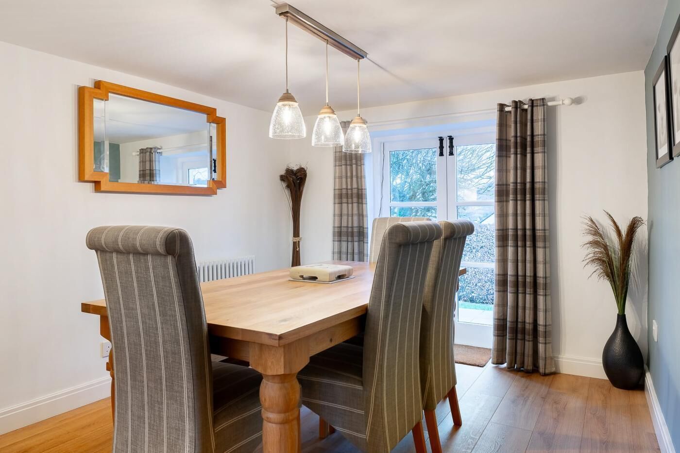Un coin repas avec une table en bois, des chaises à rayures, des luminaires suspendus, un miroir et une vue par la fenêtre.