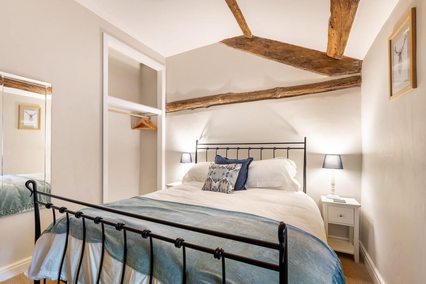 Una camera da letto con struttura del letto in metallo, trave al soffitto in legno, comodino con lampada e una cornice con un'immagine appesa al muro.