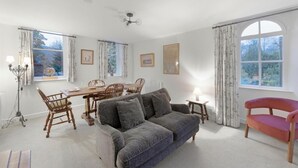 Ein geräumiges Zimmer mit Esstisch, Stühlen, einer Couch und einem roten Sessel. Es gibt Fenster mit Vorhängen, einen Deckenventilator und Wandlampen.