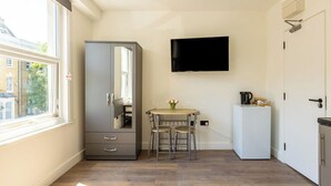 Et lille, godt oplyst værelse med et stort vindue, en grå garderobe, et lille bord med stole, et fladskærms-TV og et hvidt køleskab.