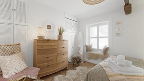 Un dormitorio con un aparador de madera, una alfombra mullida, una cama con manta blanca, un perro pequeño y una ventana con persianas.