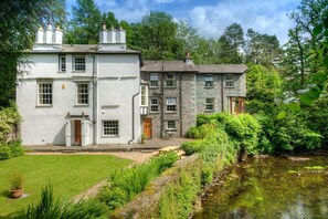 Uma casa de dois andares com fachada de pedra e exterior branco, rodeada por um jardim bem cuidado e um lago.