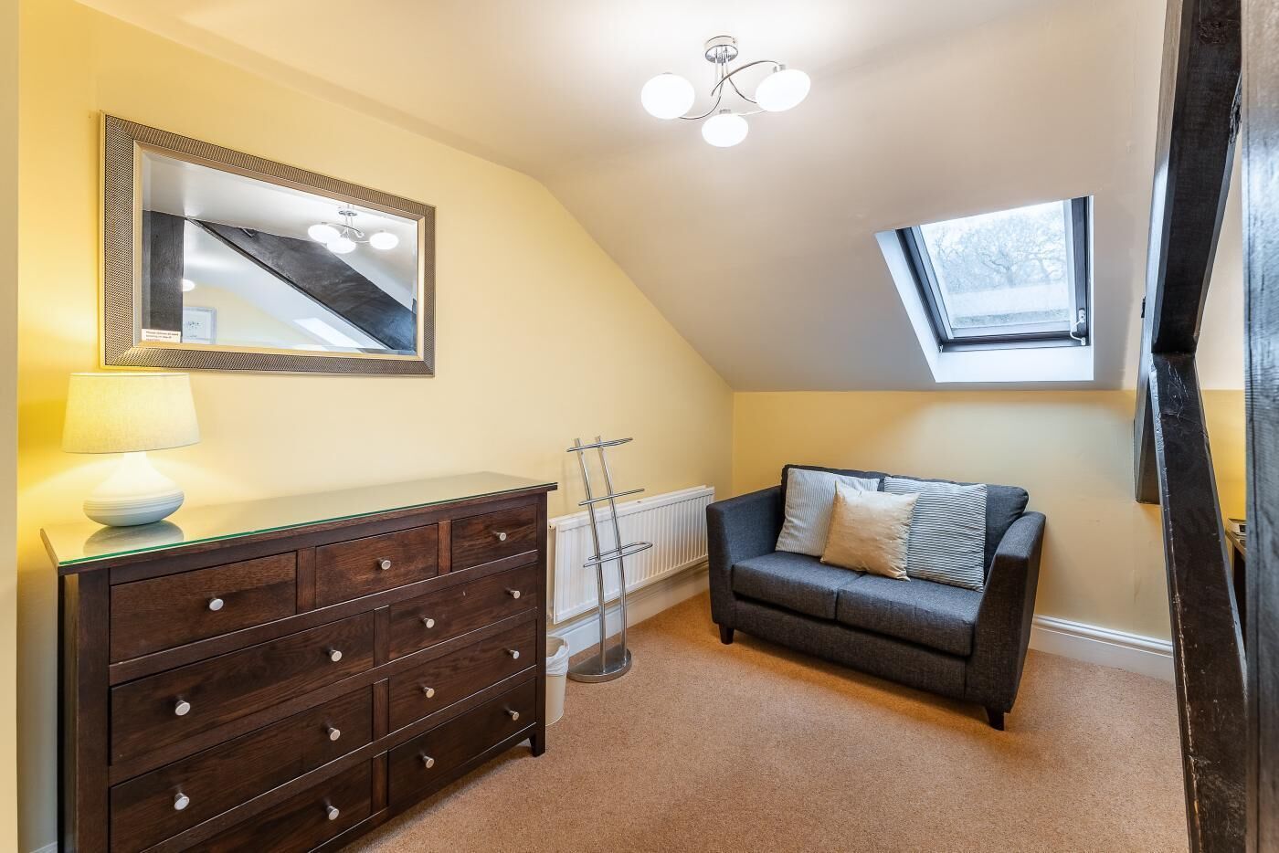A room with a dark wooden dresser, a gray sofa, a mirror, a lamp, a radiator, and a skylight.