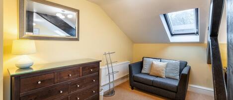 A room with a dark wooden dresser, a gray sofa, a mirror, a lamp, a radiator, and a skylight.