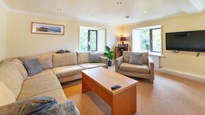A living room with a sectional sofa, a coffee table, a TV mounted on the wall, and two windows. - 4 Bed in Ambleside (oc-lr109) (Ambleside)