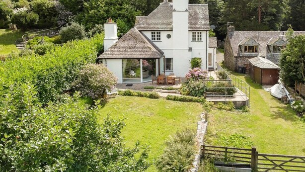 Una casa de dos pisos con un jardín amplio, un patio con mobiliario exterior y un camino de piedra que lleva a la casa.